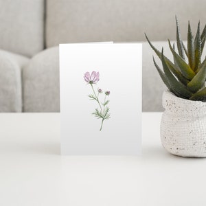 Purple cosmos flower stem watercolor notecard printed on white linen card stock with white envelope on a coffee table with an aloe plant.