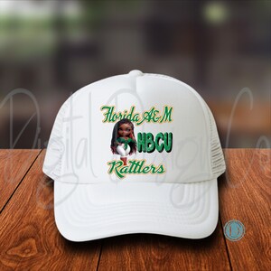 May include: White trucker hat with the text "Florida A&M HBCU Rattlers" in green and gold. The hat features a graphic of a cartoon woman. The hat is on a wooden surface.