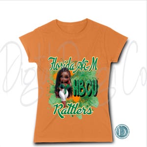 May include: Orange t-shirt featuring a graphic of a doll with long black hair, wearing a green top and white pants. The shirt has the words "Florida A&M HBCU Rattlers" in green and white script. The graphic is set against a green and orange splatter design.