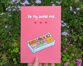 May include: A pink card with the text "to my sweet one" in white cursive writing. Three small red hearts are below the text. A cartoon illustration of a box of assorted colorful candies is below the hearts.