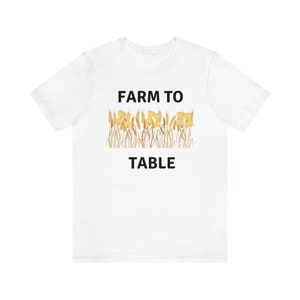 Puede incluir: Camiseta blanca con texto negro que dice "FARM TO TABLE" y un gráfico de tallos de trigo y flores amarillas.