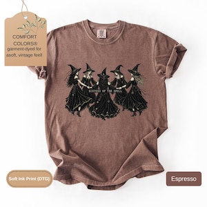 May include: An espresso-colored short-sleeve t-shirt featuring a graphic print of five figures in black dresses and pointed hats holding hands in a circle. The text "SISTERS OF THE SPELL" is printed in the center. The shirt has a crew neckline.