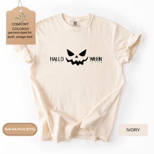 May include: An ivory short-sleeve t-shirt featuring a black graphic of a jack-o'-lantern face centered between the text "HALLO" and "WEEN" in a dripping font style. The shirt has a crew neckline and a tag indicating Comfort Colors brand.