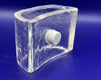 May include: A clear glass ice cube mold with a white button on the top. The mold is textured and has a curved shape.