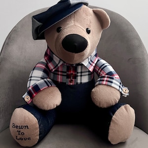 May include: A plush teddy bear wearing a blue and white checked shirt, blue denim dungarees, and a blue cap. The bear is sitting on a grey chair. The bear's foot has the text "Sewn To Love" embroidered on it.