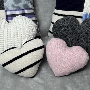 May include: Three heart-shaped knitted cushions in white, black and white stripes, and pink. The cushions are arranged on a grey surface.