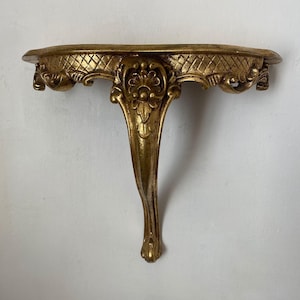 May include: A gold-colored wall shelf with ornate detailing. The shelf has a curved front edge with a diamond pattern and decorative carvings. The single leg is also gold and has a curved design.