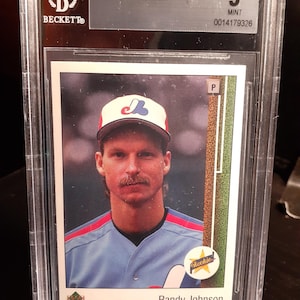 Puede incluir: Una tarjeta de béisbol Upper Deck de 1989 de Randy Johnson, calificada como 9 Mint por Beckett. La tarjeta presenta un retrato de Johnson con un uniforme de béisbol azul y rojo. La tarjeta está dentro de un soporte protector transparente.