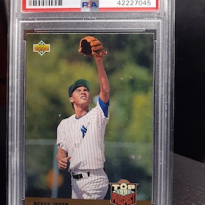 May include: A 1993 Upper Deck Derek Jeter baseball card, graded NM-MT 8 by PSA, in a protective case. The card shows Derek Jeter in a baseball uniform reaching for a ball. The text reads "1993 Upper Deck Derek Jeter".