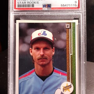 Puede incluir: Una tarjeta de béisbol Randy Johnson Star Rookie de 1989 Upper Deck, calificada como Mint 9, en un soporte protector. La tarjeta presenta a Randy Johnson con un uniforme de béisbol azul y una gorra con un logotipo. El texto de la tarjeta incluye "Rookie" y "Randy Johnson."