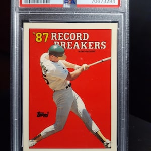 Puede incluir: Una tarjeta de béisbol de Mark McGwire de la serie Topps Tiffany de 1988. La tarjeta, protegida en un estuche, presenta un fondo rojo con el texto "'87 Record Breakers" y una imagen de McGwire en acción. La tarjeta está calificada.