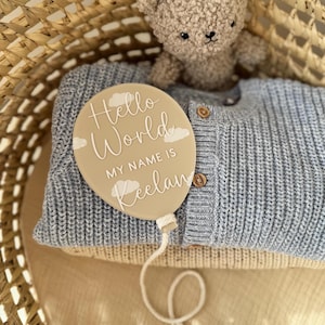 K&ouml;nnte beinhalten: Ein beigefarbenes, ballonf&ouml;rmiges Schild mit dem Text "Hello World, MY NAME IS Keelan" in Wei&szlig;, mit Wolkenakzenten. Das Schild liegt auf einem blauen Strickpullover, mit einem Teddyb&auml;r und einem Weidenkorb im Hintergrund.