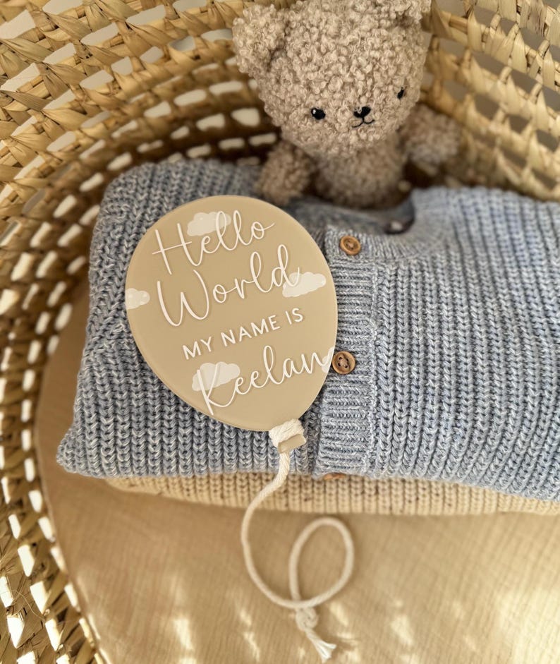 K&ouml;nnte beinhalten: Ein beigefarbenes, ballonf&ouml;rmiges Schild mit dem Text "Hello World, MY NAME IS Keelan" in Wei&szlig;, mit Wolkenakzenten. Das Schild liegt auf einem blauen Strickpullover, mit einem Teddyb&auml;r und einem Weidenkorb im Hintergrund.