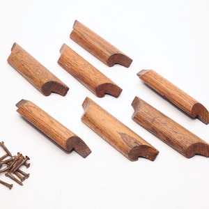 Può includere: Una collezione di sette pezzi di finitura per mobili in legno con un'estremità curva e un bordo smussato, insieme a un piccolo mucchio di viti arrugginite. Il legno ha un caldo tono marrone e un motivo a venature visibile. I pezzi sono disposti su sfondo bianco.