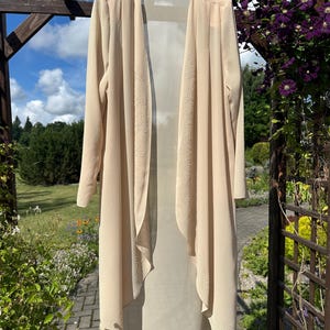 May include: A long-sleeved, open-front duster cardigan in a light beige color. The garment features delicate, tonal embroidered detailing along the front edges and the bottom hem, with a flowing, lightweight fabric construction and a straight hem.