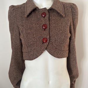 Peut inclure: Boléro marron à chevrons avec un col et trois gros boutons ronds rouges. La veste a des manches longues avec des épaules bouffantes. Le design court de la veste est visible.