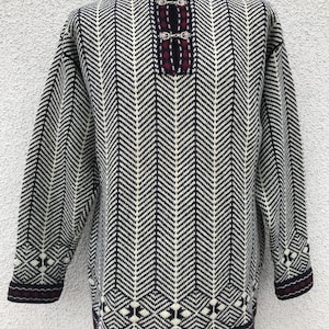 May include: A long-sleeved sweater with a black, white, and burgundy color scheme. The sweater features a herringbone pattern on the body and decorative patterns on the cuffs and hem. It has a round neckline with a burgundy trim and silver clasps.