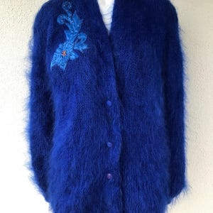 Peut inclure: Un cardigan bleu vif et duveteux avec un col en V et trois boutons. Le cardigan présente une application florale sur le côté gauche, ajoutant une touche décorative au tricot doux et texturé.