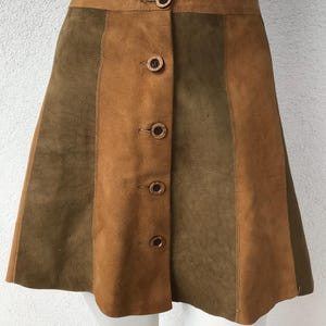 May include: A-line skirt in brown and olive green suede. The skirt has a button-up front with five round buttons and a high waist. The skirt is approximately 46 cm long.