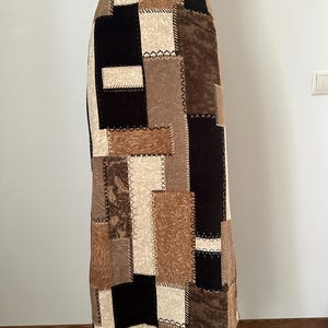 May include: A long, patchwork skirt in shades of brown, beige, and black. The skirt features a high waist and a straight silhouette. The fabric is composed of rectangular and square patches, stitched together with white thread, creating a unique, textured design.