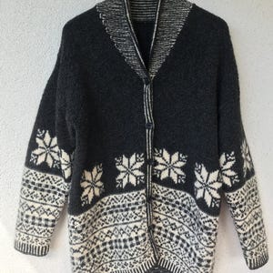 Può includere: Un cardigan grigio scuro con collo a scialle e chiusura con bottoni. Il maglione presenta un motivo a fiocchi di neve e geometrico color crema sulle maniche e sull'orlo. È appeso a una gruccia nera. Abbigliamento invernale.