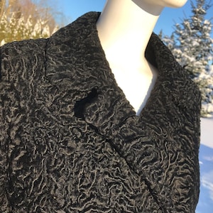 May include: Close-up of a black coat with a textured, curly surface. The coat has a wide collar and a button closure. The background shows a snowy outdoor scene with trees.