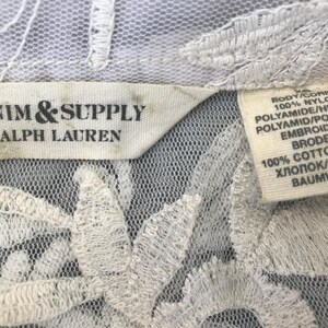 Może przedstawiać: Zbliżenie białej koronkowej tkaniny z kwiatowym haftem. Etykieta głosi "DENIM & SUPPLY RALPH LAUREN". Druga etykieta zawiera szczeg&oacute;łowe informacje o składzie tkaniny: 100% nylonu dla korpusu i 100% bawełny dla haftu.