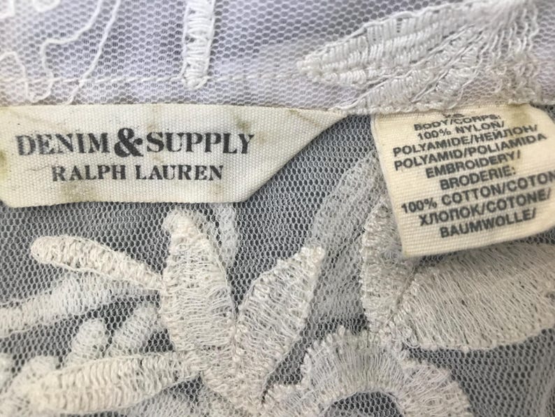 Może przedstawiać: Zbliżenie białej koronkowej tkaniny z kwiatowym haftem. Etykieta głosi "DENIM & SUPPLY RALPH LAUREN". Druga etykieta zawiera szczeg&oacute;łowe informacje o składzie tkaniny: 100% nylonu dla korpusu i 100% bawełny dla haftu.