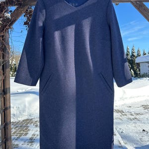 Könnte beinhalten: Ein marineblaues Kleid mit langen Ärmeln, V-Ausschnitt und zwei Vordertaschen. Das Kleid hängt an einem schwarzen Kleiderbügel vor dem Hintergrund von Schnee und blauem Himmel. Das Kleid scheint aus Wolle oder einem Wollmischgewebe zu sein.