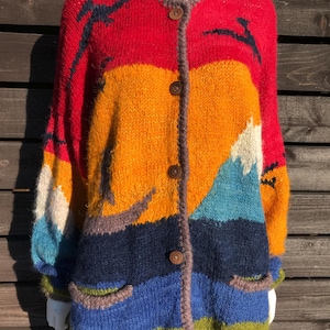 以下が含まれることがあります： カラフルなストライプ柄のボタン留めニットカーディガン。カーディガンには、山、ボート、空の風景が描かれています。