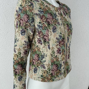 以下が含まれることがあります： 丸いネックラインと長袖のフローラルタペストリージャケット。 ジャケットは、ピンク、紫、青、緑の色合いのカラフルな花のパターンを持つベージュの背景が特徴です。 フルレングスのジッパーが付いています。