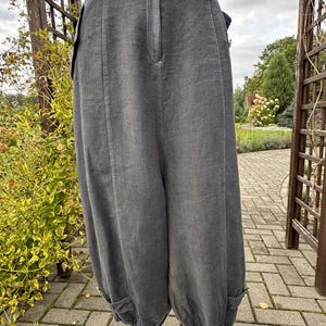 May include: Charcoal gray trousers featuring a high-waisted design with a front button closure, vertical seam detailing, and side pockets. The legs have a relaxed, wide-cut silhouette that tapers toward the hem, which includes small buttoned tabs.