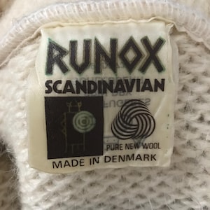 May include: Close-up of a tag from a Runox Scandinavian garment. The tag is rectangular with the word "RUNOX" in green letters. Below it, "SCANDINAVIAN" is printed. The tag also features a logo and states "PURE NEW WOOL MADE IN DENMARK".