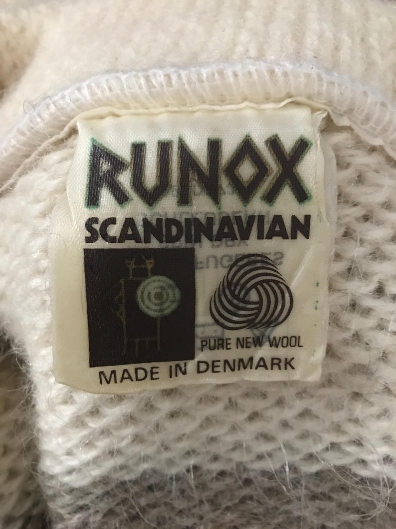 May include: Close-up of a tag from a Runox Scandinavian garment. The tag is rectangular with the word "RUNOX" in green letters. Below it, "SCANDINAVIAN" is printed. The tag also features a logo and states "PURE NEW WOOL MADE IN DENMARK".