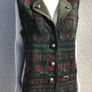 Könnte beinhalten: Eine grüne, rote und braune gemusterte ärmellose Weste mit einem grünen Cordkragen und silbernen Knöpfen. Die Weste hat eine Tasche auf der linken Seite und ein silbernes Metalletikett auf der rechten Seite.