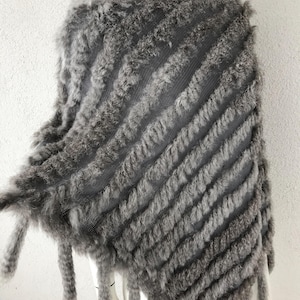 May include: A grey, textured poncho with a diagonal striped pattern. The poncho is made of a soft, furry material and has fringe along the bottom edge. The garment is displayed on a white mannequin.