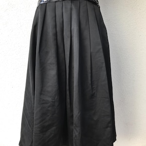 May include: A black skirt with a wide, sequined waistband. The skirt is pleated and falls to the mid-calf. The waistband features a bow detail. The skirt is made of a smooth, shiny fabric.