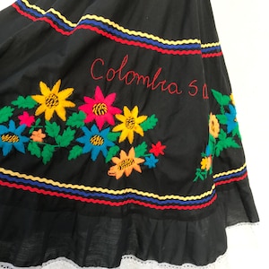 以下が含まれることがあります： 黒いスカート。カラフルな刺繍の花と赤い「Colombia」の文字が特徴。スカートの裾には白いレースのトリムがあり、赤、黄、青の水平バンドがあります。伝統的な衣装です。