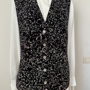 Puede incluir: Un chaleco de terciopelo negro con un estampado moteado y botones plateados. Se lleva sobre una camisa blanca con cuello. El chaleco tiene cuello en V y un corte entallado, adecuado para ocasiones formales o semiformales.