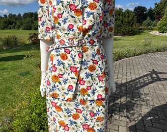 Op de afbeelding: Een tweedelige outfit bestaande uit een top met korte mouwen en knoopsluiting en een bijpassende rok, beide voorzien van een levendig bloemenpatroon met rode, oranje, gele en blauwe bloemen op een witte achtergrond, inclusief een stoffen riem.