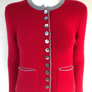 Peut inclure: Veste rouge en tricot avec une bordure grise autour du col, des poches et de la boutonnière. La veste a un col rond, des manches longues et une rangée de boutons sur le devant. Deux poches sont visibles.