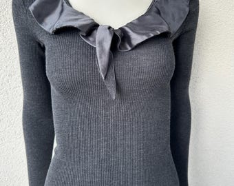 May include: A charcoal gray long-sleeve ribbed knit top featuring a wide, rounded neckline adorned with a smooth, satin-like ruffled collar. The collar includes a small, tied fabric detail at the center front, and the hem and cuffs have a subtle, wavy finish.