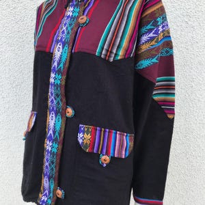 Könnte beinhalten: Schwarze Kapuzenjacke mit farbenfrohen gewebten Einsätzen und Akzenten. Die Jacke hat eine Knopfleiste vorne, zwei Vordertaschen und einen Kordelzug am Saum. Die gewebten Details umfassen geometrische und florale Muster in Lila, Türkis und Orange.