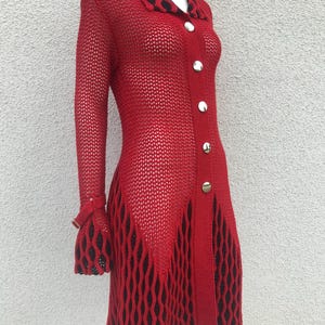 Puede incluir: Vestido rojo de tejido abierto, con mangas largas y cuello. El vestido presenta botones plateados en la parte delantera y detalles negros en forma de diamante en el dobladillo y los puños. El vestido se exhibe en un maniquí.