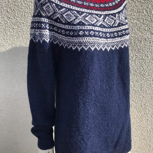 Puede incluir: Un suéter azul marino con cuello rojo y patrones geométricos blancos. El suéter de manga larga tiene cuello redondo y se muestra en un maniquí. El diseño presenta una combinación de colores lisos y detalles de punto intrincados.