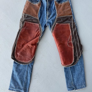 Peut inclure: Jean bleu avec des empiècements en cuir marron et brun rougeâtre cousus sur le devant. Les empiècements en cuir recouvrent l'avant des jambes et des poches. Le jean a une conception classique à cinq poches.