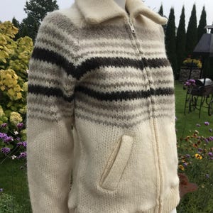 May include: A cream-colored zip-up sweater with horizontal stripes in shades of brown and gray. The sweater has a collared neckline, a front pocket, and ribbed cuffs and hem. The garment is displayed on a mannequin.