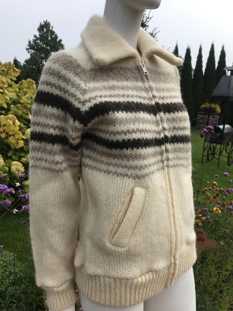 May include: A cream-colored zip-up sweater with horizontal stripes in shades of brown and gray. The sweater has a collared neckline, a front pocket, and ribbed cuffs and hem. The garment is displayed on a mannequin.