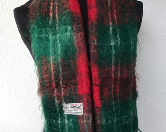 May include: A green, red, and brown tartan scarf with a fringed edge. The scarf is displayed on a black mannequin. A small tag with text is visible on the scarf.