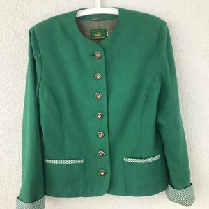 Puede incluir: Una chaqueta verde con cuello redondo y botones dorados. La chaqueta tiene dos bolsillos delanteros con ribete a cuadros blancos y verdes. Los puños también están ribeteados con el mismo patrón de cuadros. La chaqueta está colgada de una percha blanca.